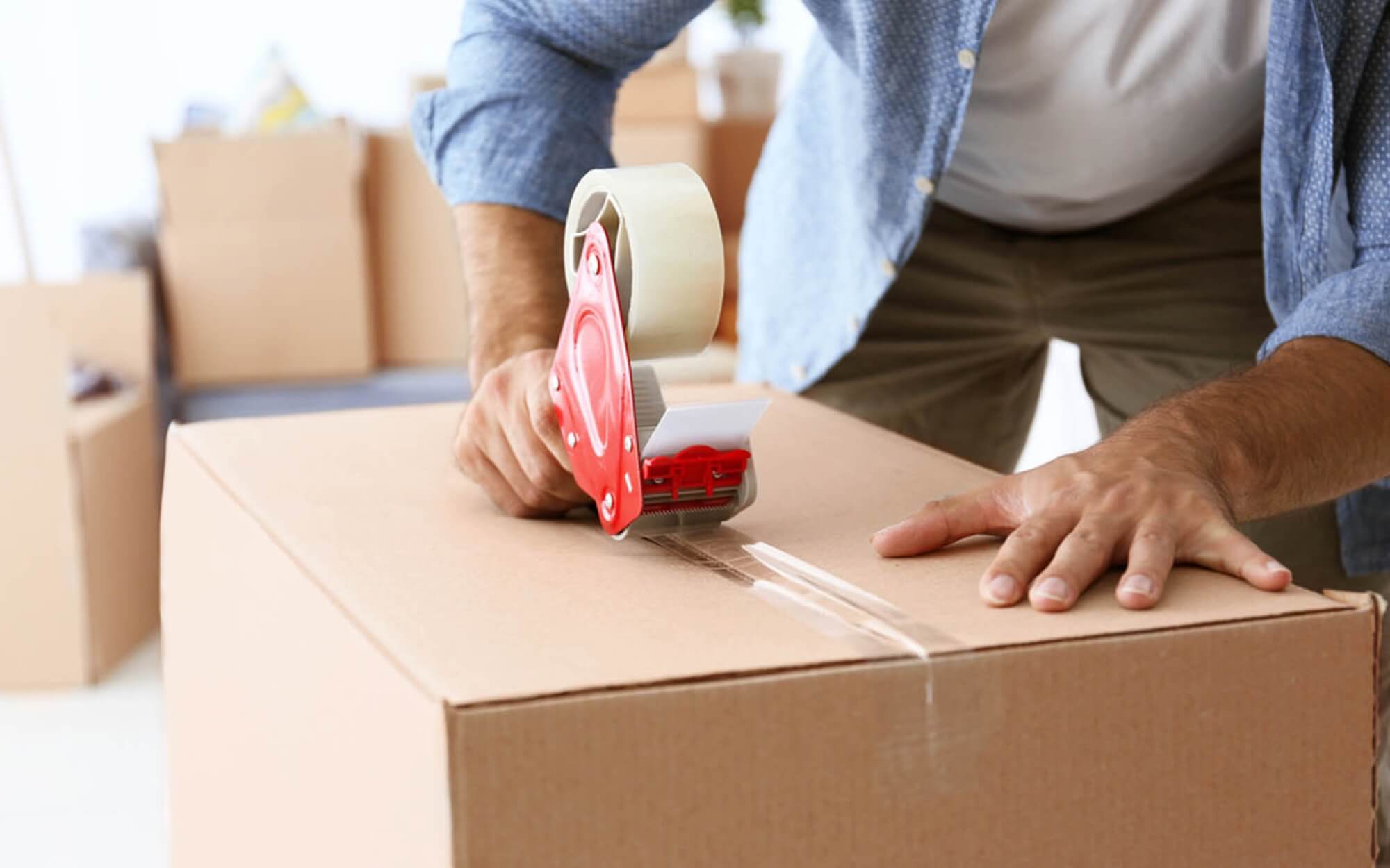 A person seals a moving box with packing tape in a room filled with boxes for moving.